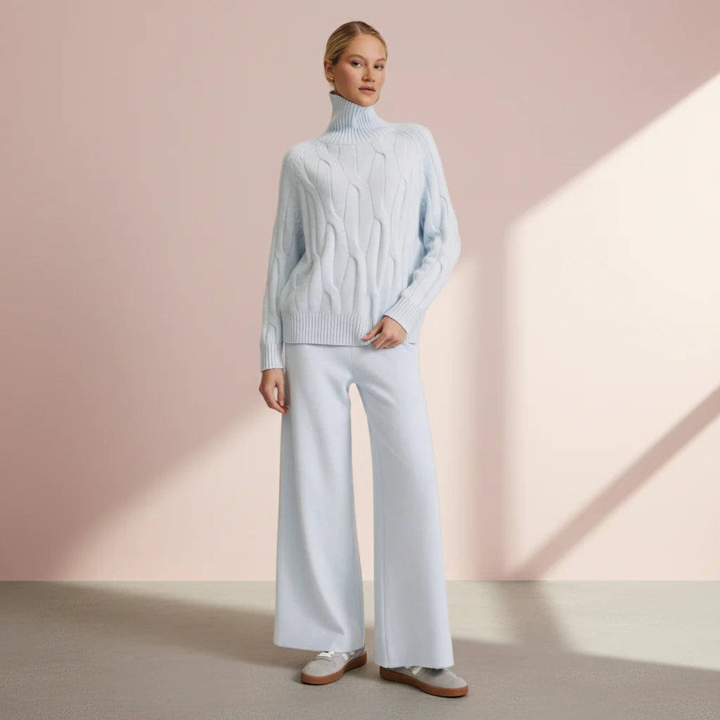 Woman wearing a light blue cable knit sweater and white pants against a pink background