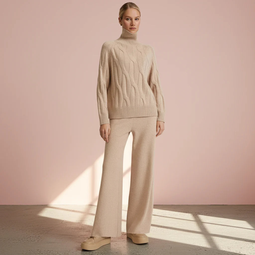 Woman wearing a beige cable knit sweater and pants against a pink wall.