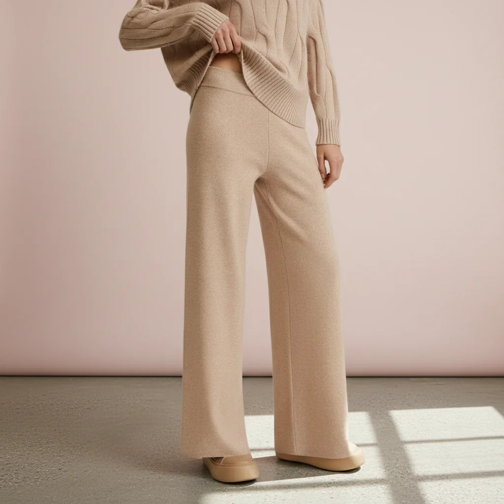 Person wearing a beige sweater and wide-leg pants against a pink wall.