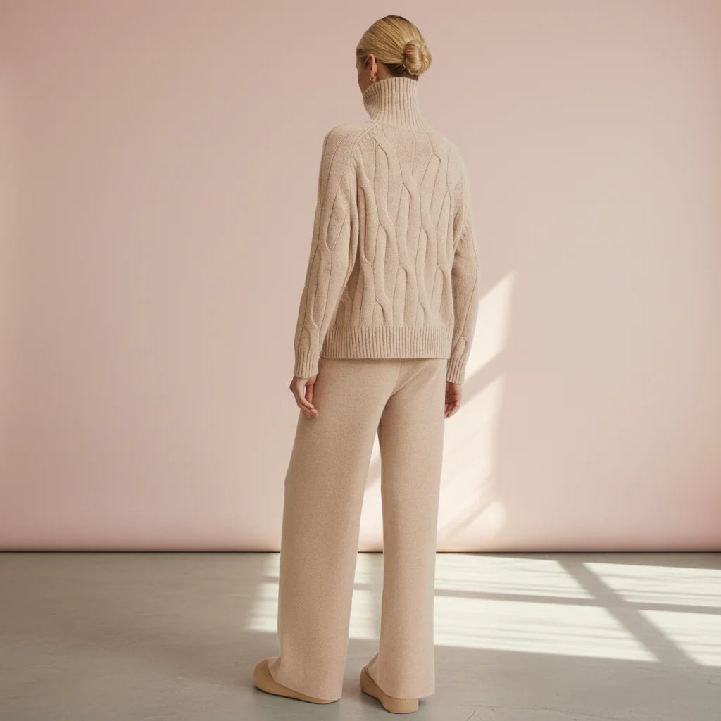 Person wearing a beige cable knit sweater and pants against a pink wall.