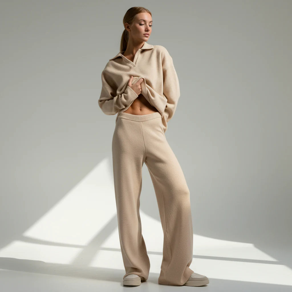 Woman wearing a beige sweater and pants standing in a well-lit room with a soft light source.