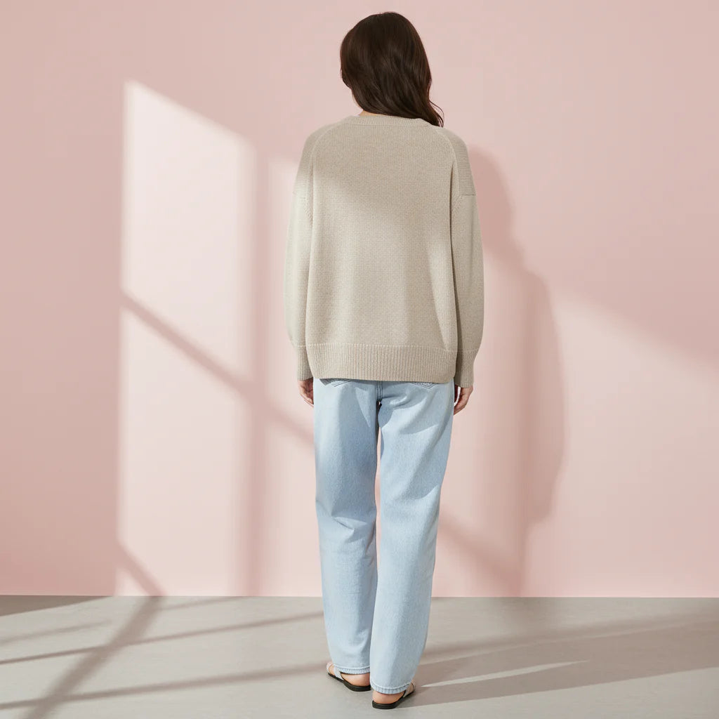 Person wearing a beige sweater and light blue jeans against a pink wall.