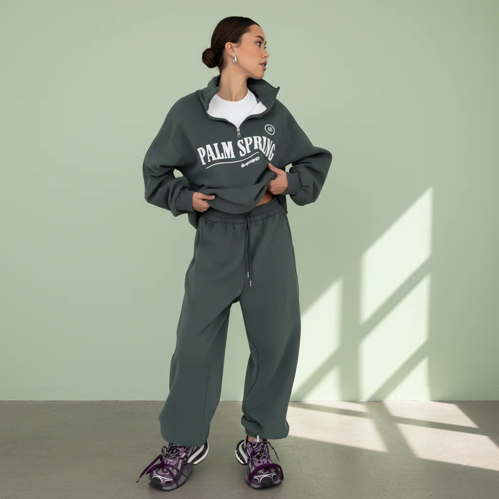 Person wearing a gray 'Palm Spring' tracksuit against a light green wall.